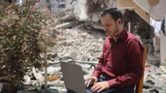 رجل فلسطيني يعمل على حاسوب محمول في الهواء الطلق، محاطاً بالأنقاض في غزة.
