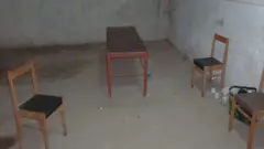 Una habitación desnuda, con paredes y suelo de hormigón. En ella hay tres sillas de madera y una pequeña mesa de madera, con unas cuantas botellas de agua de plástico en el suelo.