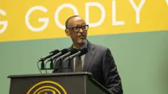 Perezida w'u Rwanda Paul Kagame arimo kuvuga ijambo avugira mu ndangururamajwi enye ziri ku meza maremare yo kuvugiraho ijambo, ariho ikirango cya Repubulika y'u Rwanda. Yambaye indorerwamo, ikositimu yijimye ijya kuba ubururu, karuvati y'ikigina, n'ishati ijya kuba ubururu bwerurutse. Arareba ahagana ku ruhande. Inyuma he hari ikirango cy'icyatsi kibisi n'umuhondo, cyanditseho mu mweru mu cyapa ngo 'GODLY', andi magambo ntaboneka ku ifoto. Ifoto yo ku cyumweru, i Kigali, ku itariki ya mbere Gashyantare (2) mu mwaka wa 2026. 