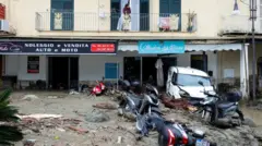 A woman looks from a balcony at a street covered in debris and damaged vehicles