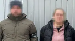 двоє людей із заблюреними обличчями, один із них чоловіків у куртці хакі, співробітник СБУ, а жінка в рожевій кофті на замочку та в чорній куртці - затримана і підозрювана в злочині