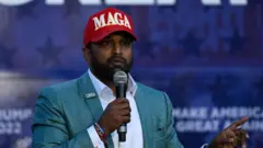 Kash Patel, wearing a red MAGA hat, and speaking into a microphone and pointing