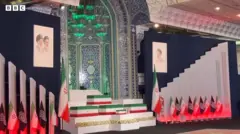 イラン国旗の緑・白・赤3色の棺が複数並び、前最高指導者ハメネイ師らの肖像画が掲げられている