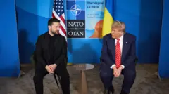 Volodimir Zelenszkij ukrán elnök (bal oldalon) és Donald Trump amerikai elnök (jobb oldalon) a hágai NATO-csúcson, 2025. június 25-én. A két politikus egy kis asztal két oldalán ül, Zelenszkij feketébe öltözve, kicsit Trump felé fordul. Trumpon sötétkék öltöny van, fehér ing és piros nyakkendő fehér csíkokkal, az öltönyén kis amerikai zászló kitűző. A háttérben látható a NATO/OTAN kétnyelvű felirat.

