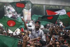 Um jovem com uma faixa na cabeça exibindo a bandeira de Bangladesh agita uma grande bandeira do país e grita palavras de ordem. A bandeira é verde-escura, com um círculo vermelho no centro. Dezenas de manifestantes o cercam, a maioria usando faixas idênticas, durante ato do movimento estudantil, que marcou um mês desde a destituição da ex-primeira-ministra de Bangladesh, Sheikh Hasina, em Daca, Bangladesh, em 5/9/2024