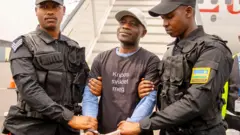 François Gasana cyangwa Franky Dusabe uri hagati yambaye ingofero y'umweru n'umupira w'umukara afashwe n'abapolisi babiri ku mpande zombi