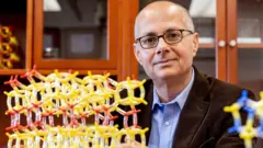 UC Berkeley chemistry professor Omar Yaghi, wearing a blue shirt, brown jacket and glasses, poses in his lab at the University of California in Berkeley US. Next to him is a model showing metal-organic frameworks made of yellow and red tubes and nodes.