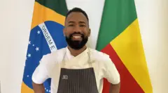 Homem com avental de cozinheiro sorrindo em frente a bandeiras do Brasil e Benin