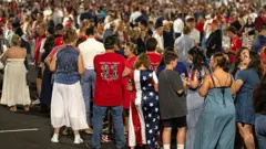 Um grande grupo de pessoas faz fila para entrar no Estádio State Farm em Glendale, Arizona, EUA. Uma mulher usa um vestido estampado com as estrelas e listras da bandeira dos Estados Unidos. Outro homem, de costas, veste uma camiseta vermelha com o número 31 e os dizeres: "Obrigado, Charlie".