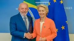 Foto de Lula e da presidente da Comissão Europeia, Ursula von der Leyen. Ele usa um terno azul marinho claro e ela usa um conjunto laranja. Ambos estão de mãos dadas e diante de bandeiras da União Europeia e do Brasil.