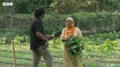 সবজি খেতের মধ্যে হাতে শাক নিয়ে দাঁড়িয়ে থাকা একজন নারীর সঙ্গে কথা বলছেন বিবিসি বাংলার সাংবাদিক