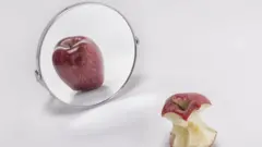 Uma maçã comida em frente a seu reflexo no espelho, onde aparece inteira