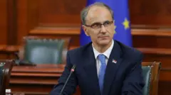 Ђуро Мацут у тегет оделу у скупштиној сали, а иза њега је застава Европске уније