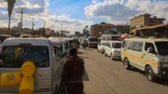 wasu motoci a kan hanya, inda aka ɗaura wa wata jarka uku , wani mutum ya juya bayansa a jikin wata mota