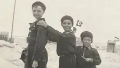 Três crianças vestidas para o inverno em uma paisagem nevada. A criança do meio apoia as mãos no ombro da criança mais alta, à esquerda, que sorri. A criança menor, à direita, também sorri. Construções cobertas de neve e duas bandeiras, incluindo uma com a suástica, são visíveis ao fundo, sugerindo um cenário da época da Segunda Guerra Mundial.