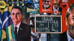 Um homem está em pé ao lado de uma barraca de rua que vende toalhas dos candidatos à presidência Luís Inácio Lula da Silva e Jair Bolsonaro na Avenida Paulista, em 23 de setembro de 2022, em São Paulo, Brasil. A barraca exibe uma placa com o número de toalhas de cada candidato vendidas até o momento.
