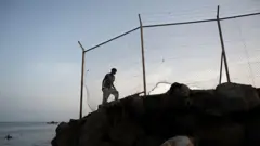 Fas ile İspanya'nın Kuzey Afrika'daki özerk şehri Ceuta arasındaki sınırda bir göçmen, kayalıkların üzerindeki tel örgülerin yanında yürüyor. Arkada deniz görünüyor.