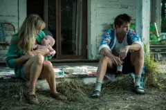 En una escena de la película Die My Love, Jennifer Lawrence carga a un bebé mientras está sentada en una acera. A su izquierda, indiferente, Robert Pattinson sostiene entre sus manos una cerveza.