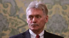 Dmitry Peskov é um homem branco, com bigode e cabelos grandes. Ele usa um terno preto, gravata vermelha e blusa branca