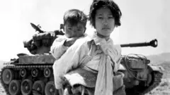 Mulher com criança, com tanque de guerra ao fundo