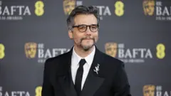 O ator Wagner Moura posando no tapete vermelho do Bafta. Ele veste um terno preto clássico com lapela acetinada, camisa branca e gravata preta. Usa óculos de armação escura e tem barba e cabelos grisalhos bem aparados. Na lapela, há um broche prateado em formato floral. Ao fundo, aparece o painel oficial do evento com o logotipo do BAFTA e a máscara dourada característica da premiação.