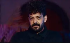 الليثي شعره أسود ولحية سوداء قصيرة، يرتدي قميصاً أسود ووراءه خلفية بتدرجات اللون الخمري والبنفسجي.