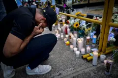 Un hombre con una rodilla en el suelo llora frente a flores y velas encendidas en el suelo