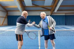 Dois jogadores de tênis idosos se cumprimentam junto à rede da quadra