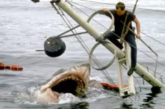 No set de gravação de 'Tubarão', o ator Roy Scheider (1932-2008) aparece sobre os restos de um barco que afunda, enquanto o tubarão o espera com a boca aberta na água