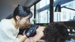 A tatuadora Gong Greem com o cabelo preso para trás, aplicando um estêncil no braço e nas costas de uma cliente