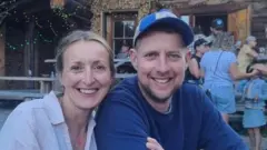 Caroline veste uma camisa clara e Gareth um moletom azul escuro. Ambos estão sorrindo. Ao fundo, é possível ver um pequeno grupo de pessoas, incluindo uma criança