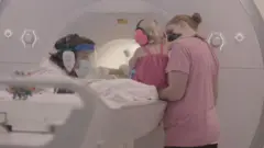 Um bebê usando protetores auriculares sentado ao lado de uma máquina de ressonância magnética funcional, segurado pela mãe, com o pesquisador Turk-Browne do outro lado.
