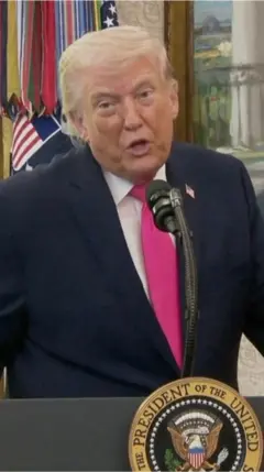 කළු කෝට් එකක්, සුදු පැහැති කමිසයක් සහ රෝස පාට ටයි එකක් පැළඳ සිටින ඇමෙරිකානු ජනාධිපති ඩොනල්ඩ් ට්‍රම්ප් මයික්‍රපෝනයක් ඉදිරිපිට කතා කරන ආකාරය දැක්වේ.