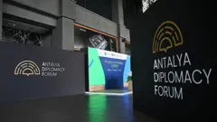 Üzerinde İngilizce Antalya Diplomasi Forumu yazan dekorların olduğu bir fotoğraf. Arkadaki bir panoda üse "Tüm gözler Antalya'da" başlığıyla Kasım'da düzenlenecek COP31 İklim Zirvesi'ne dair bilgi yer alıyor.
