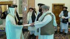 راست سفیر قطر که لباس مخصوص عربی سفید بر تن دارد و در مقابلش رئیس الوزرای حکومت طالبان که لباس سفید، واسکت روشن، لنگی نقره‌ای و دستمال سفید رنگ بر شانه دارد در حال گرفتن اعتمادنامه سفیر قطر است. آنطرف چند مقام دیگر طالبان ایستاده اند.