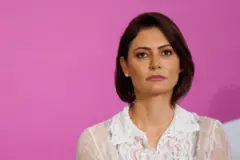 Michelle Bolsonaro olha para a frente com uma expressão séria. Ela tem cabelos curtos castanhos escuros e veste uma camisa branca rendada transparente e um brinco de pérola. O fundo atrás dela é rosa