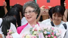 Чхве Мал Джа в розовом пиджаке и очках, с короткими поседевшими волосами, улыбается и держит в руках букеты цветов. Вокруг нее улыбающиеся молодые женщины