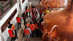 Pessoas em uma cervejaria. Elas usam coletes laranjas