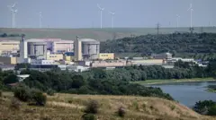 川沿いの緑地の中に原子力発電所が立ち、原子炉を覆う灰色の円柱が二つ並んでいる。背後の丘陵には風力発電の風車が並んでいる