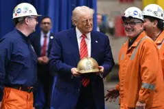 Trump segurando um capacete dourado com o nome dele ao lado de trabalhadores que usam macacões laranjas e capacetes brancos