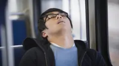 Homem asiático dorme com cabeça inclinada quando está sentando em um transporte público. Ele usa óculos, uma camisa azul e um casaco preto.