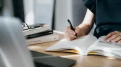 Uma pessoa segurando uma caneta sobre um livro numa mesa.