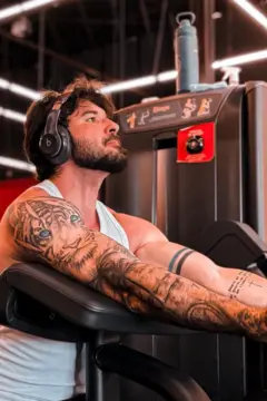 A imagem mostra um homem sentado em um aparelho de musculação em uma academia, apoiando os braços no equipamento enquanto olha para cima. Ele veste uma regata branca, usa fones de ouvido e tem os braços cobertos por tatuagens, incluindo o desenho de um tigre no braço esquerdo. Ao fundo, aparecem equipamentos de treino e a iluminação em LED característica do ambiente da academia.