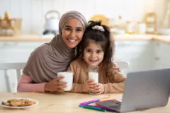 امرأة محجبة تجلس بجانب طفلة على طاولة في مطبخ، تمسك كل واحدة منهما كوب حليب، أمامهما حاسوب محمول، وقطع من البسكويت (الكوكيز) في صحن أبيض. 