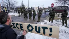 Un manifestante con un cartel que dice "¡Ice out!", "¡Fuera Ice!", parado frente a una fila de agentes federales migratorios