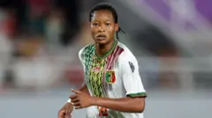 Agueissa Diarra, vêtue d'un maillot blanc du Mali orné d'un motif coloré vert, jaune et rouge sur le devant, regarde vers la caméra alors qu'on la voit courir pendant un match de football, filmée à partir de la taille.