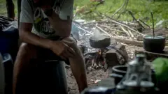 Imagem de panela e fogão improvisado. Um homem não identificado está sentado ao lado dela