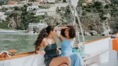 Dos mujeres visiblemente relajadas y felices, disfrutan de un paseo en barco por el mar Mediterráneo. Al fondo se divisa la famosa vista de Positano, Italia.