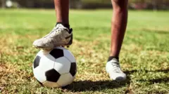 Gros plan sur les jambes d'un garçon, dont un pied repose sur un ballon de football.
