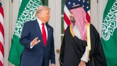 دونالد ترامپ و محمد بن سلمان در کنار هم ایستاده و در حال گفت‌وگو هستند، پرچم‌های دو کشور آمریکا و عربستان سعودی در پشت سر آنها